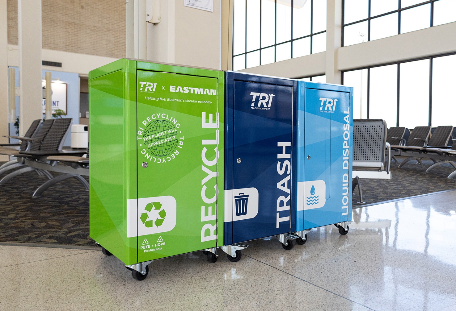 A recycling receptacle in an airport terminal is green and is marked Tri Recycling in white letters. 